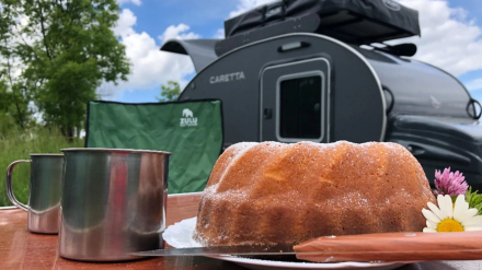 Mesa de camping con bizcocho casero y minicaravana Caretta de fondo durante una ruta gastronómica por España.